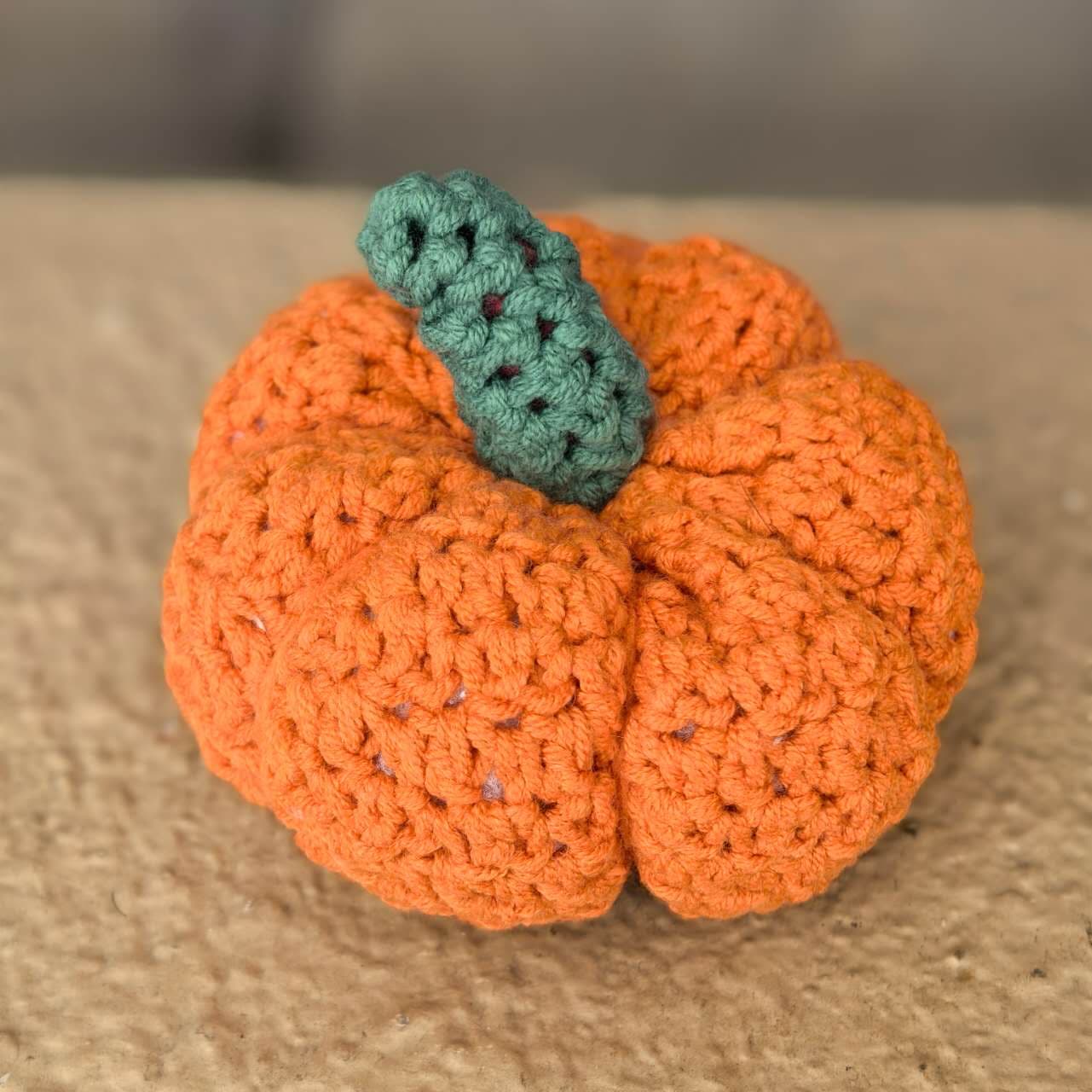 A small crocheted pumpkin in bright orange, its body pulled into seven deep vertical segments, with a chunky sage-green stem on top, resting on a stone surface in daylight.