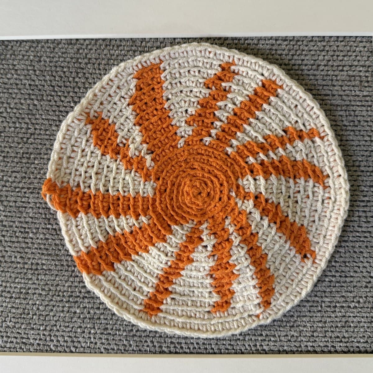 Un medallón tejido en crochet tapestry de dos colores: un molinete naranja estilo estallido de estrella sobre un disco crema, montado en un marco blanco.