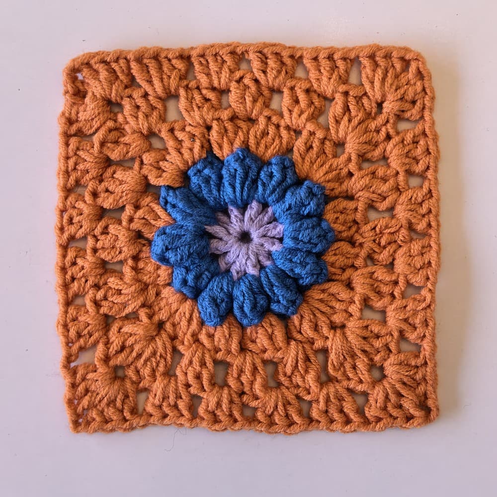 Un cuadrado de la abuela tejido a ganchillo con una flor popcorn azul en relieve y un centro lila sobre fondo terracota.